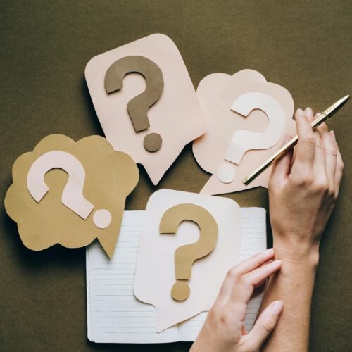 Hands holding pen next to paper question marks on notebook, symbolizing inquiry and creativity.
